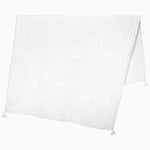 The Velvet White Throw by John Robshaw, hand quilted by skilled Indian artisans, drapes elegantly over the surface showcasing its neatly folded form, plain white hue, subtle grid pattern, and small tasseled corners. - 31272448557102