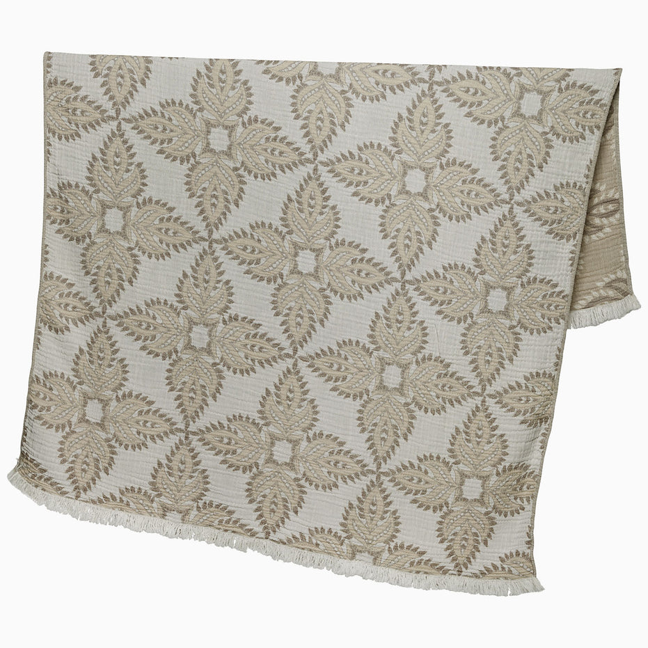 A John Robshaw Dasati Sand Throw, featuring a beige fabric with an intricate leaf-like woven design and fringed edging, draped open against a plain background.