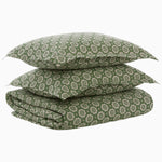 A stack of two green patterned pillows and a folded matching blanket from the Atulya Moss Duvet Set by John Robshaw, featuring a celestial design bedding motif against a plain background. - 31272398225454