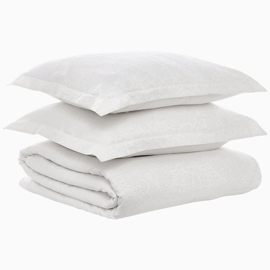 A neatly folded Amina Ecru Duvet Set by John Robshaw and two stacked white pillows with a subtle floral pattern rest on a plain white background, showcasing the finest quality cotton linen.