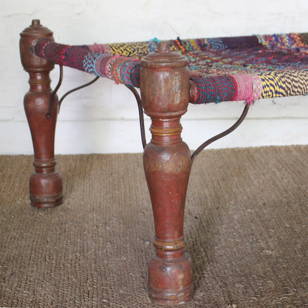Hand-turned teak legs of the John Robshaw Rangana Teak Charpoy rest atop a textured rug, while vibrant multicolored threads are woven across its frame.