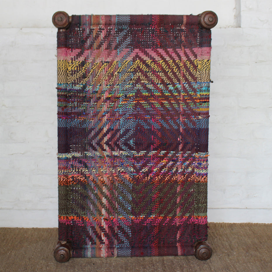 The Rangana Teak Charpoy by John Robshaw is a rectangular woven piece featuring multicolored geometric patterns crafted from vibrant threads, displayed vertically on four turned teak wooden legs against a white brick wall.