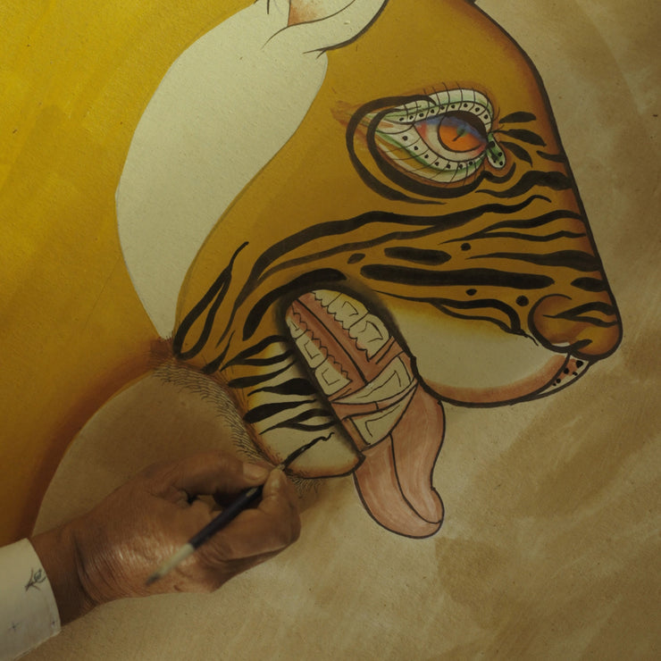 A person paints details on the face of a stylized tiger artwork, focusing on the mouth and tongue, with visible brush and hand.