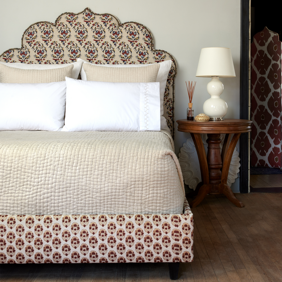 A bed with a patterned headboard covered in cotton chambray coverlets and adorned with hand stitching, featuring the Vivada Sand Woven Quilt by John Robshaw.