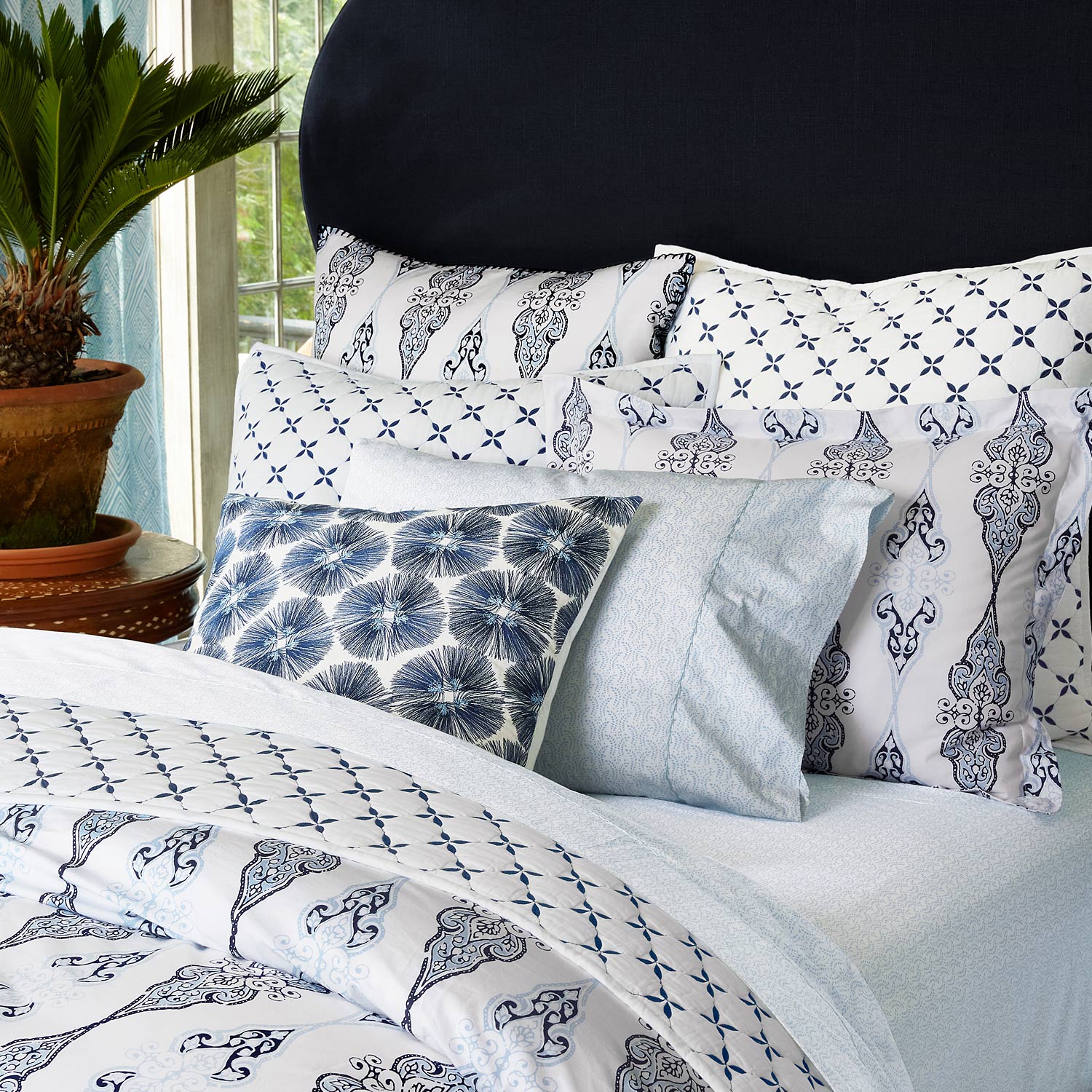 A Layla Indigo Quilt by John Robshaw adorns a bed with blue and white bedding, accompanied by a potted plant, reminiscent of Mughal gardens.